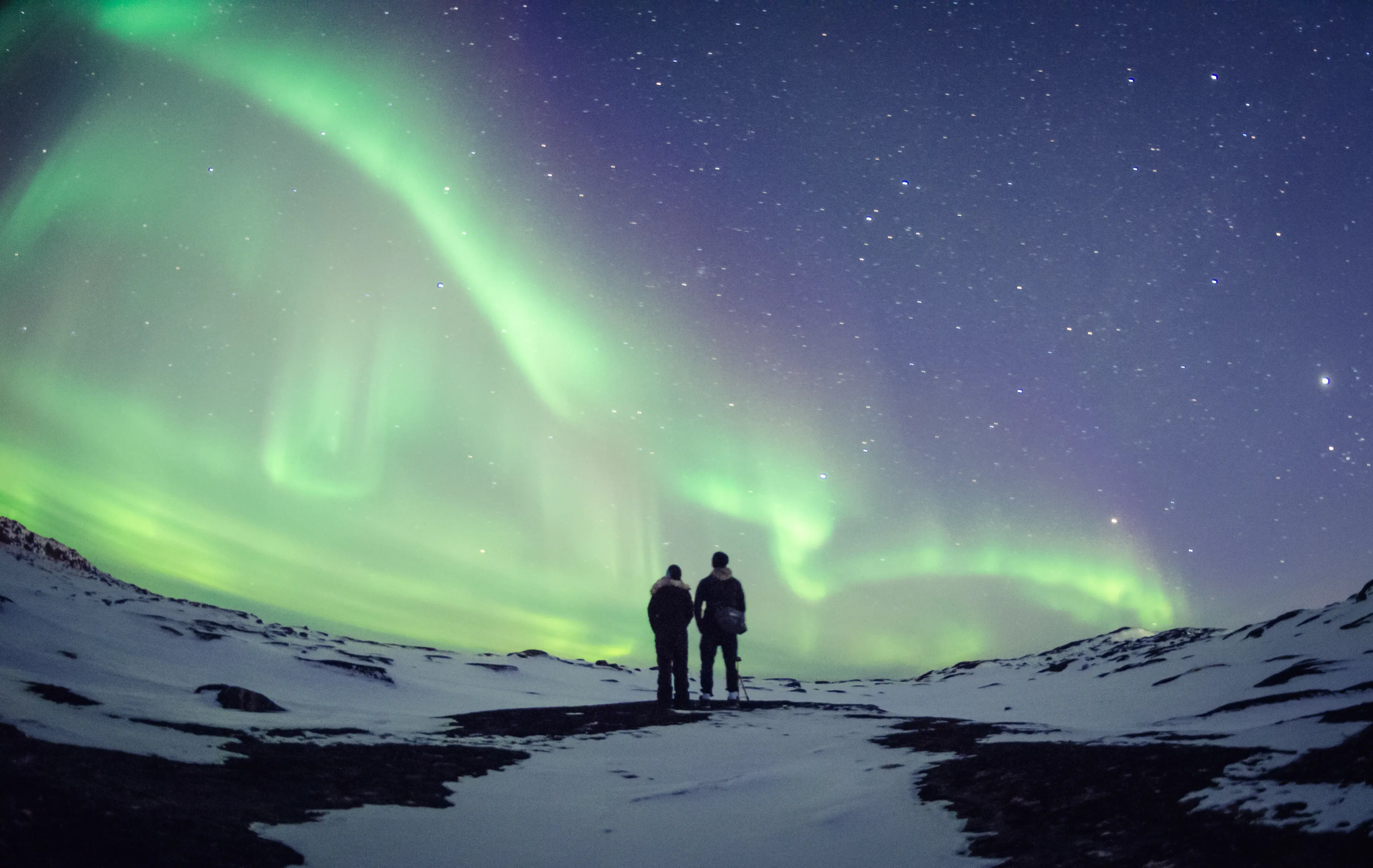 Two people on a snowy hill admiring the Aurora Borealis at night